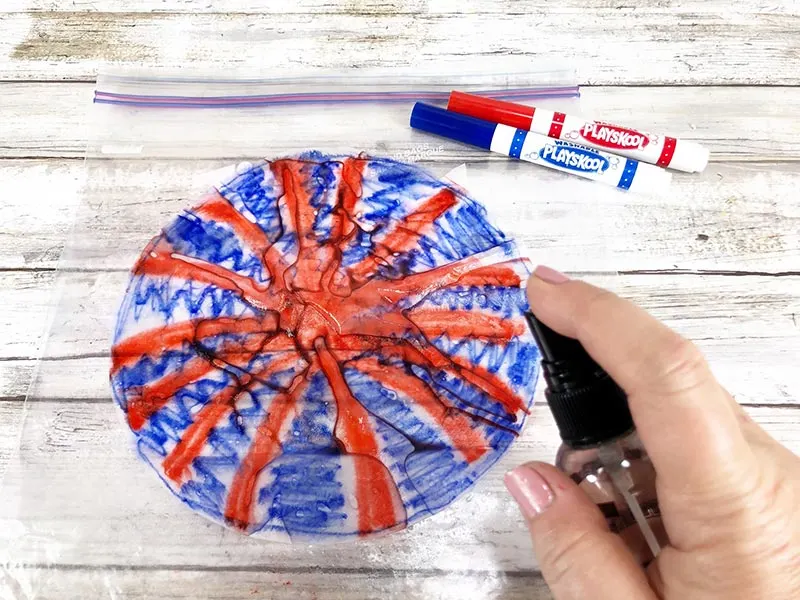 White woman's hand using spray bottle to spritz red and blue colored coffee filter with water. Filter is laying on a plastic ziptop bag.