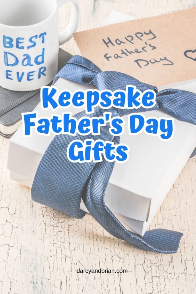 Blue text outlined with white says Keepsake Father's Day Gifts in the center over a background with a white gift box wrapped with a blue necktie. A mug with "Best Dad Ever" is also in the background and a card envelope that says Happy Father's Day.