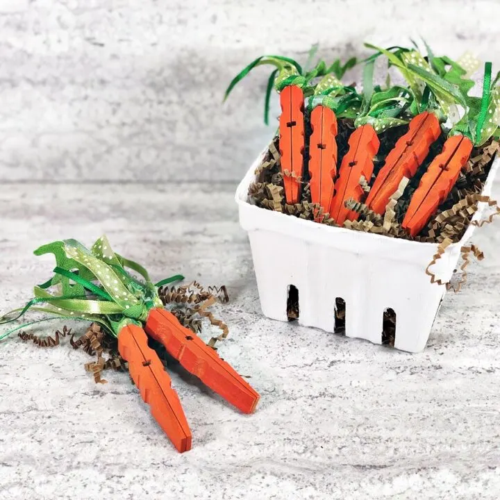 5 completed clothespin carrots laying on top of brown paper shred in a small white box made to look like a vegetable carton. Two more carrots lay on the table next to the box.