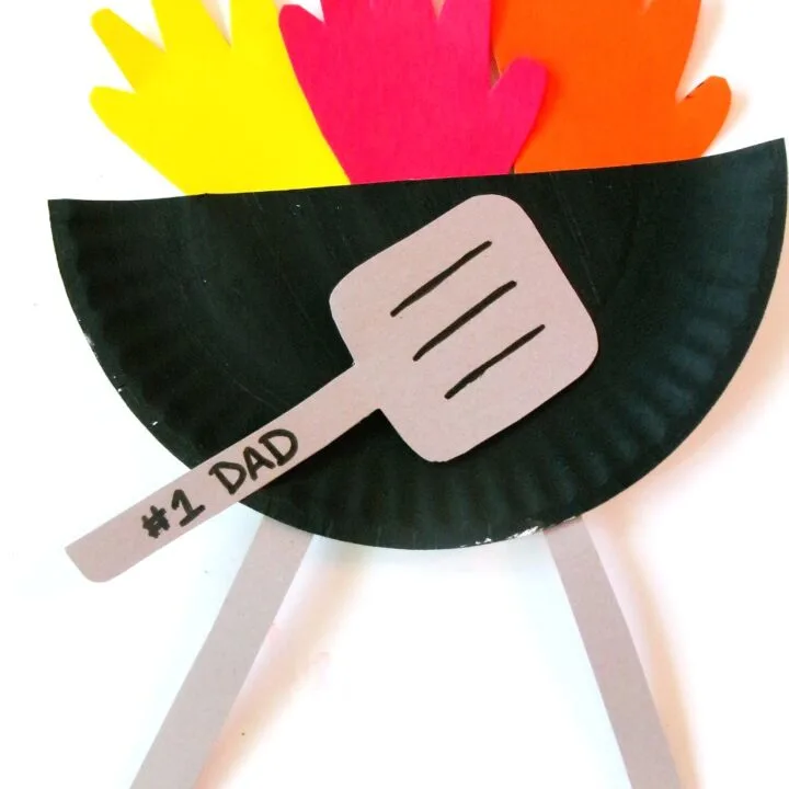 Finished craft with grill made out of black paper plate, paper handprint flames, and gray paper spatula with #1 Dad written on the handle.