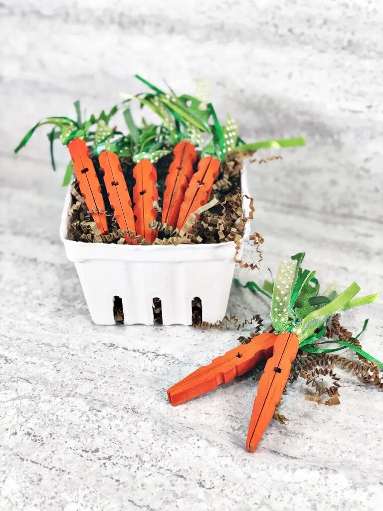 Cute carrot clothespins laying in a painted veggie box and a couple laying on the table on top of brown shredded paper.