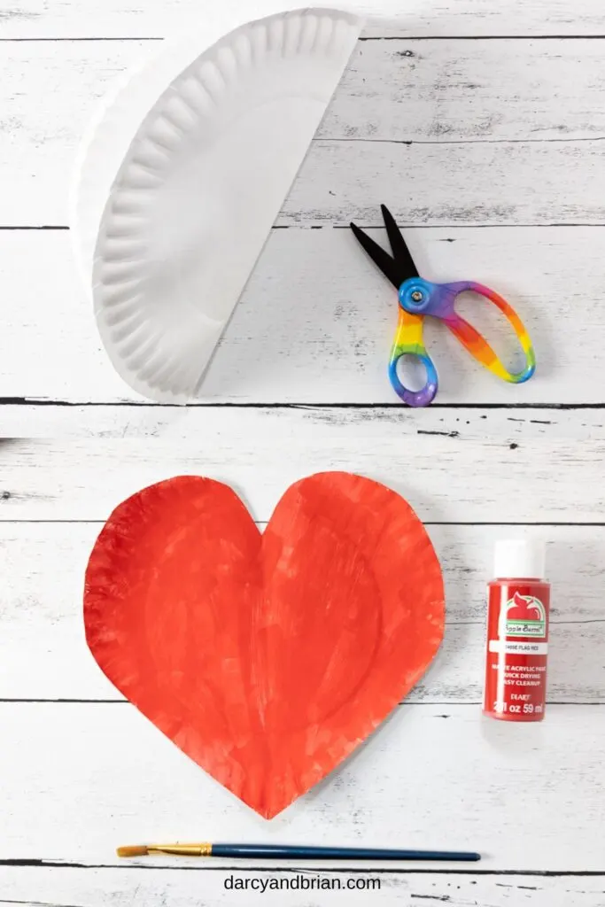 Top image shows a white paper plate folded in half next to a pair of scissors. Bottom image shows the plate in a heart shape, painted red. Bottle of paint is next to it. Paintbrush laying underneath.