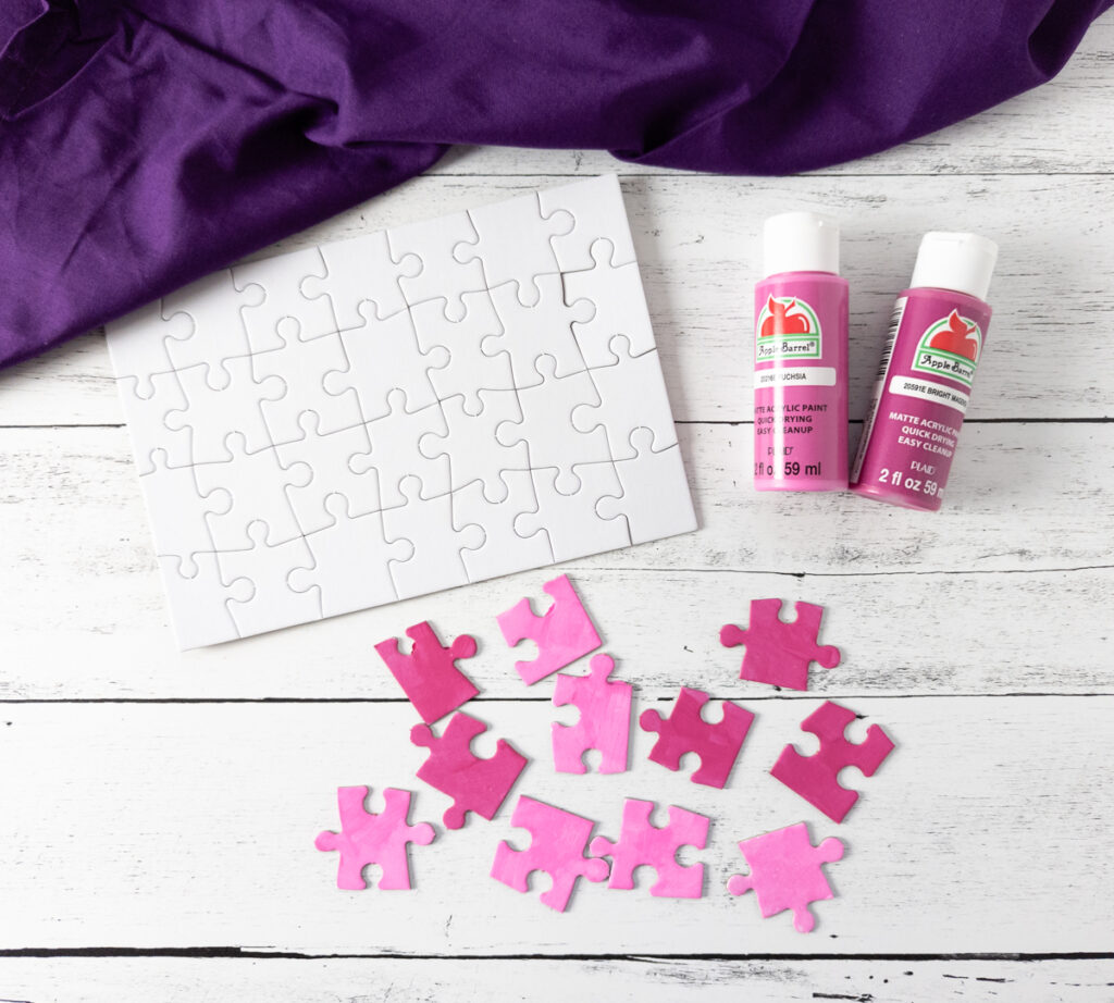 Dark purple fabric along the top. Blank puzzle with pieces connected next to two paint bottles: fuchsia and magenta. Below that are assorted puzzle pieces painted with the two different colors.