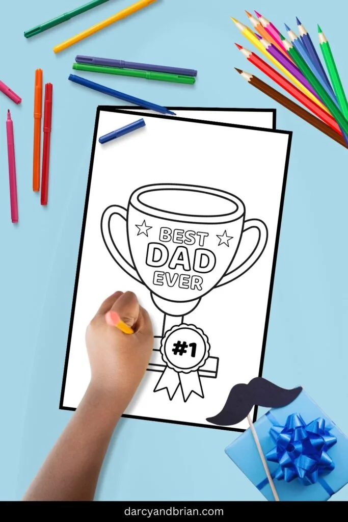 Child's hand holding a pencil over a mockup image of the trophy Father's Day card. Colored pencils scattered along the top. Bottom right corner has a fake mustache and a gift box with a blue bow on top.