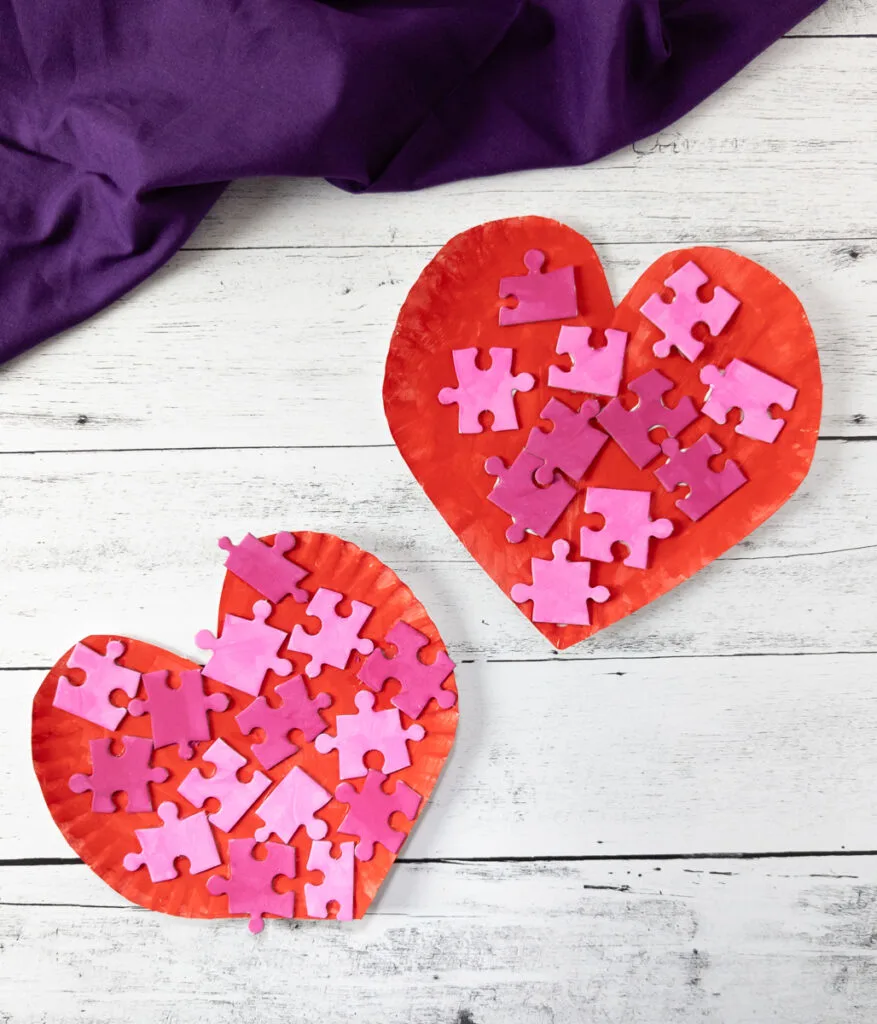 Two paper plates cut into heart shapes with painted puzzle pieces glued to them.