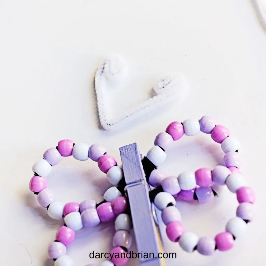 Pink, purple, and white beaded pipe cleaner wings clipped in a light purple clothespin. White chenille stem bent and curled to look like antennae laying above the project.