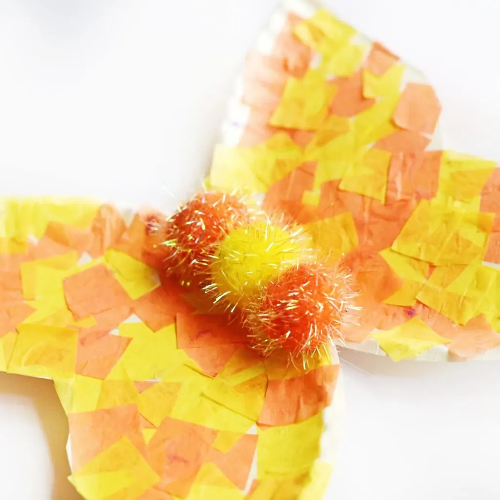 Close up showing three glittery pom poms glued in a row in center of plates to make butterfly body. Two orange pom poms with a yellow in the center.
