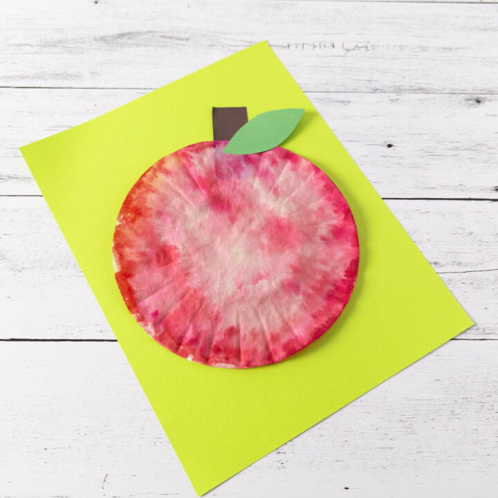 Top down angled view of finished craft to make an apple using a coffee filter. The red apple is laying on bright green paper.