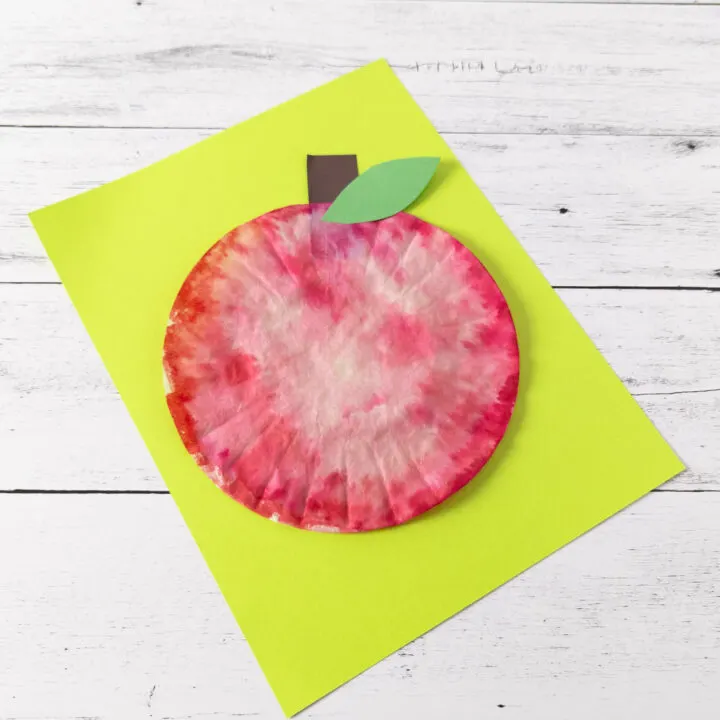 Top down angled view of finished craft to make an apple using a coffee filter. The red apple is laying on bright green paper.