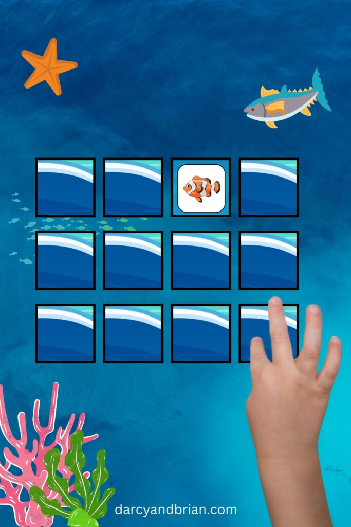 A set of cards with a blue water background flipped face down in a grid. One card face up showing a clownfish. A child's hand pointing at another card.
