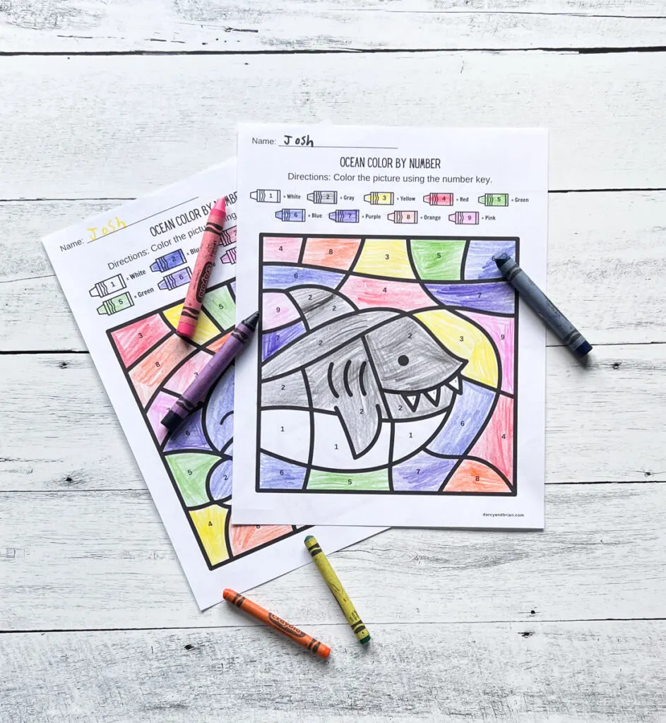 Cute shark preschool coloring by numbers worksheet filled in and crayons laying around.