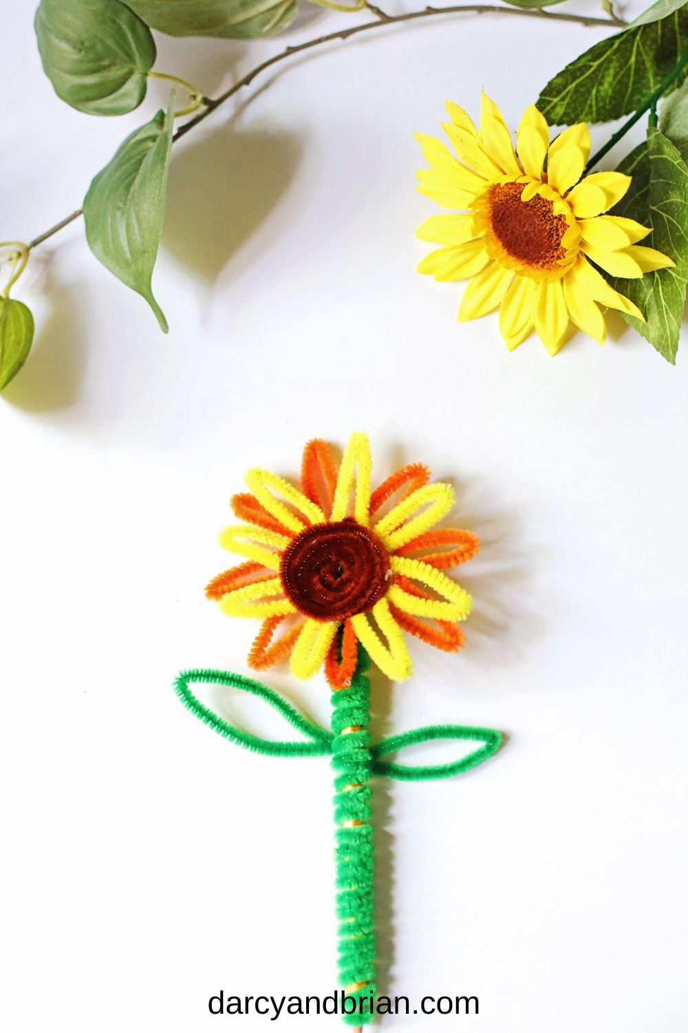 Sunflower made with pipe cleaners on a pencil laying on a white table. An artificial sunflower lays across the top of the photo.