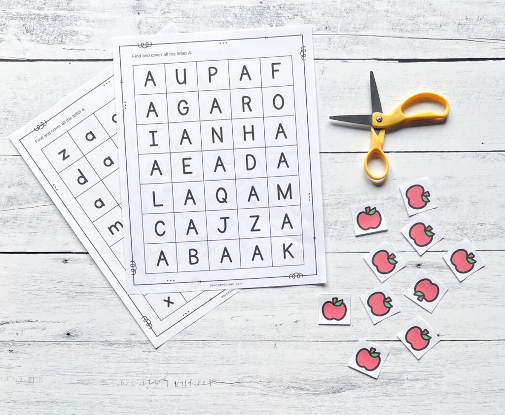 Letter A search activity pages overlapping each other on a white wood background. Several small paper apples lay nearby with a pair of scissors.