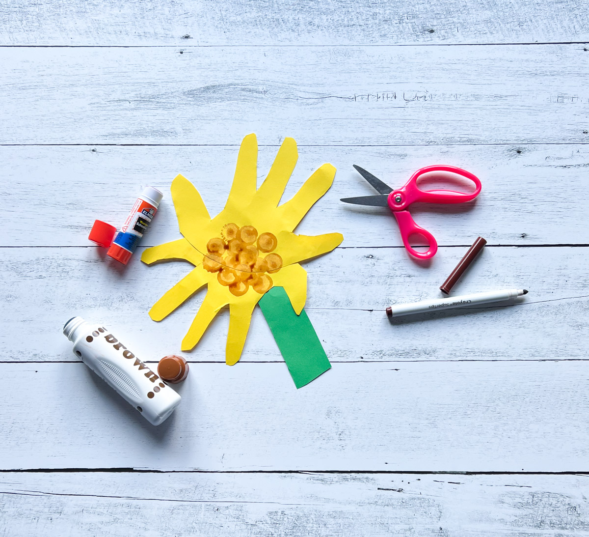 An example of a paper sunflower made with handprint petals and a short, thick stem. markers, glue stick and scissors laying next to it.