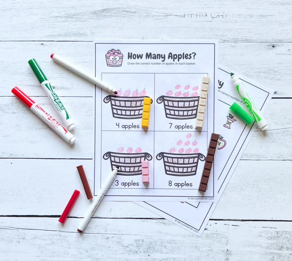 Worksheet with baskets where kids draw the number of apples to match the number provided. Worksheet is completed. Markers lay around paper.