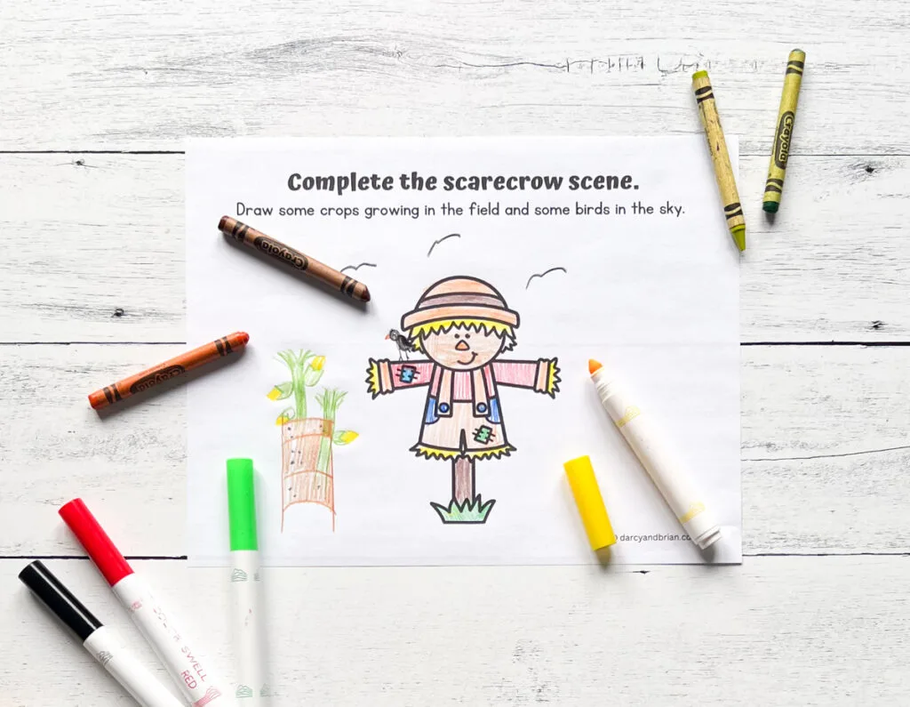 Drawing activity page to complete the scene. Scarecrow is colored in and corn and birds are drawn around it. Crayons and markers lay by the paper.