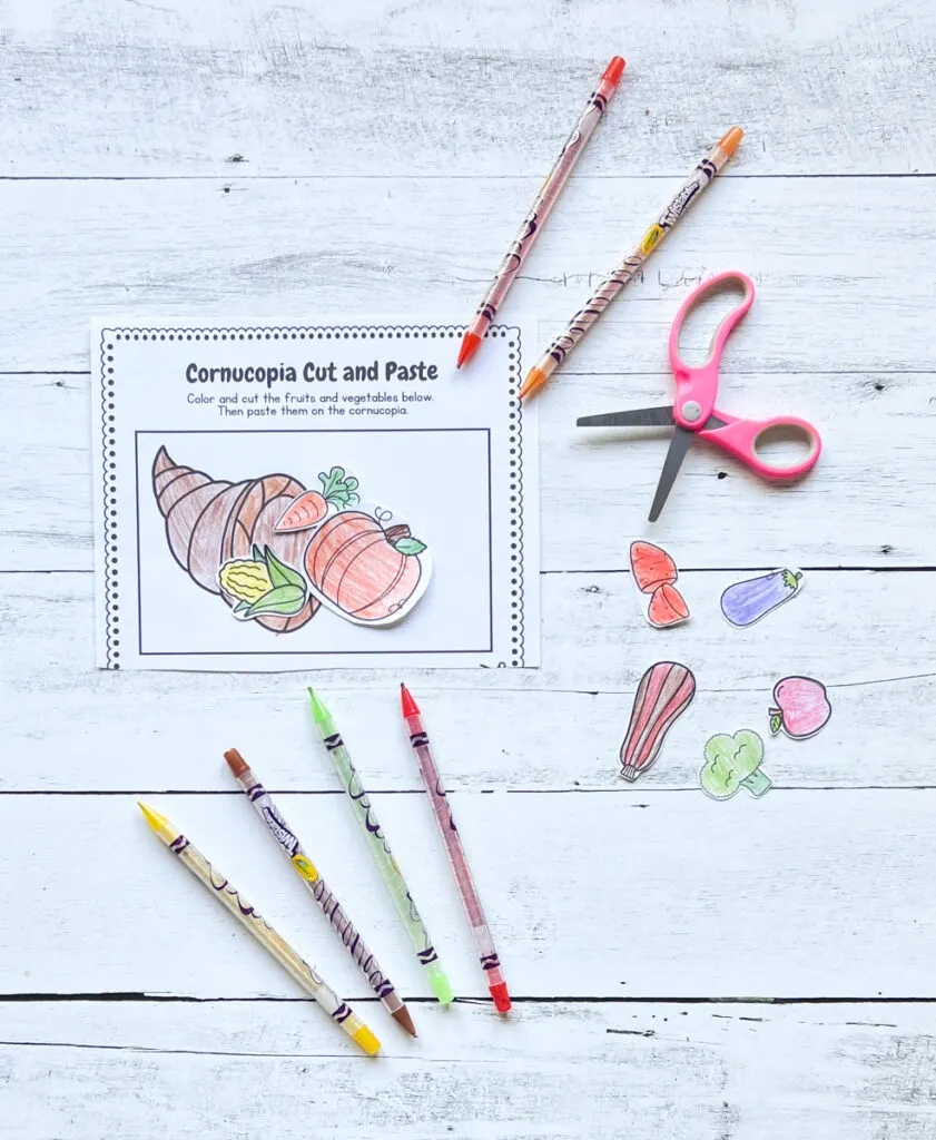 There's a cornucopia ready sample worksheet with some materials like the color pens, scissors, and colored fruits and vegetables placed on a black and white wood background.