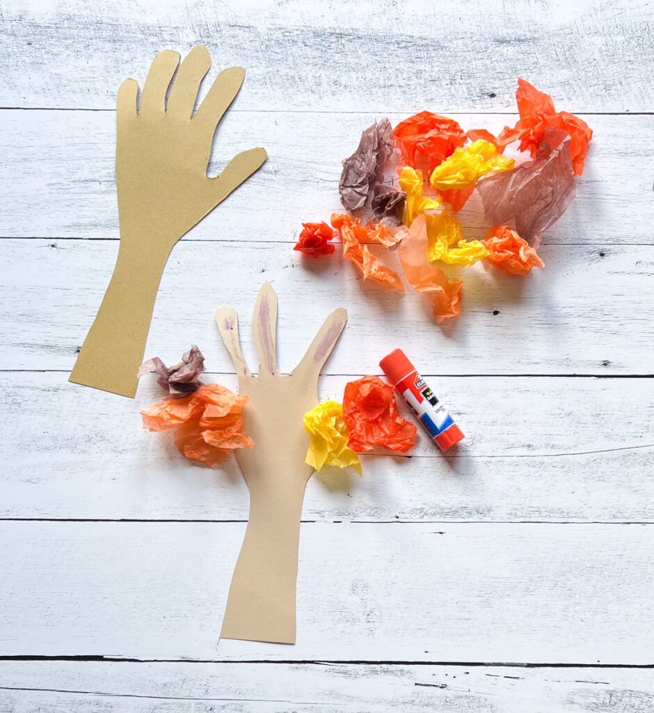 There are crumpled tissue paper with fall colors, a glue stick, and paper hands. All placed in a black and white wood background.