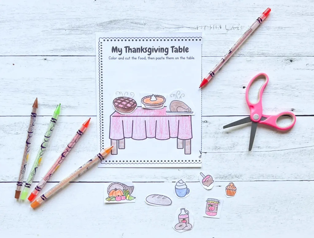 There's a page of the worksheet, color pens, scissors, cut food ready to be pasted on the Thanksgiving Table placed in a black and white wood background.