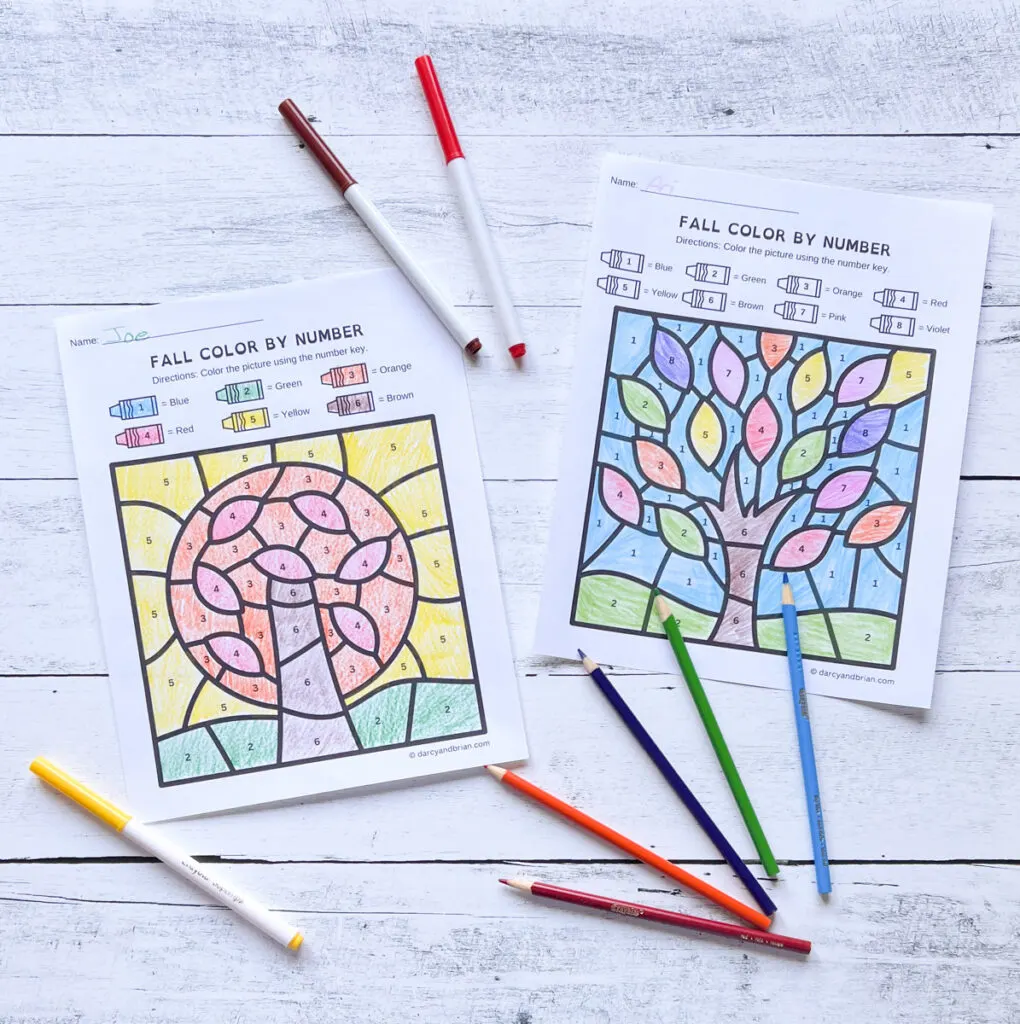 Two different color by number fall tree worksheets filled in. Colored pencils and markers lay next to the papers.