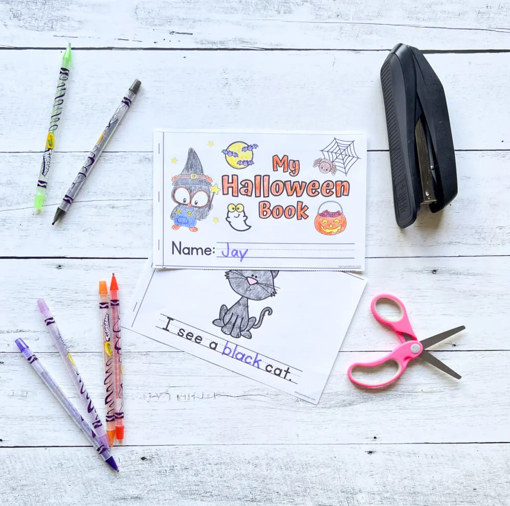 The cover of the Halloween printable book is colored in and overlapping a page with a black cat. Crayons, stapler, and scissors lay next to the papers.