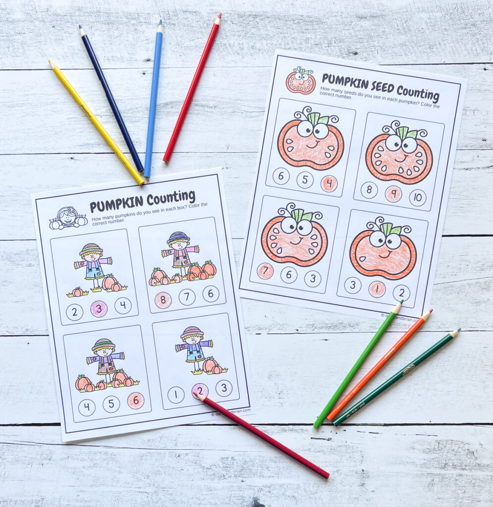 Two preschool math worksheets, one with pumpkins by scarecrows and the other counting pumpkin seeds. The pictures are colored in and the number answer is filled in. Colored pencils lay near the pages.