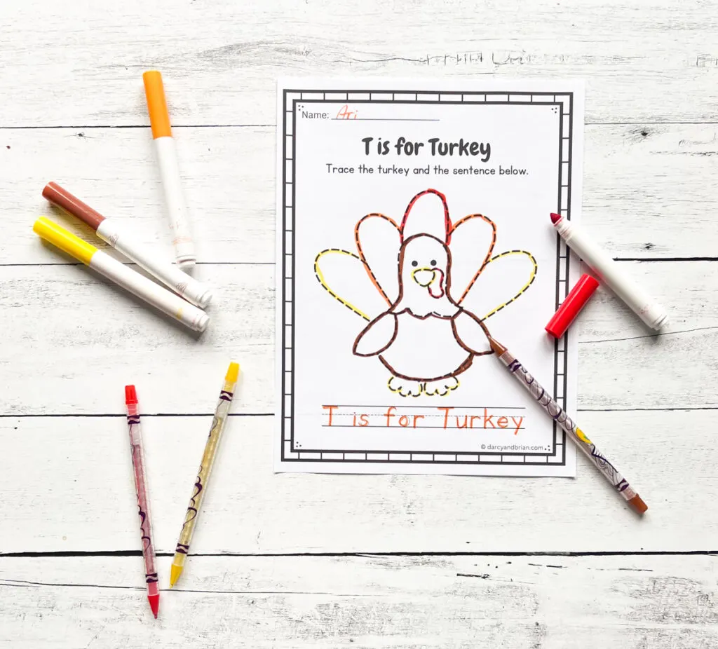 A picture of a turkey traced with red, orange, yellow, and brown markers. The sentence T is for Turkey is also traced in orange. Markers and twistable crayons lay around the paper.