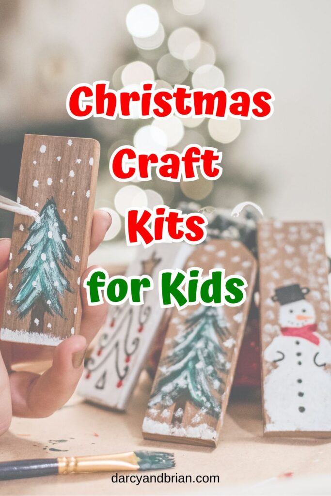 Red and green text outlined with white says Christmas Craft Kits for Kids over a background with small pieces of wood painted with snowmen and snowy pine trees.