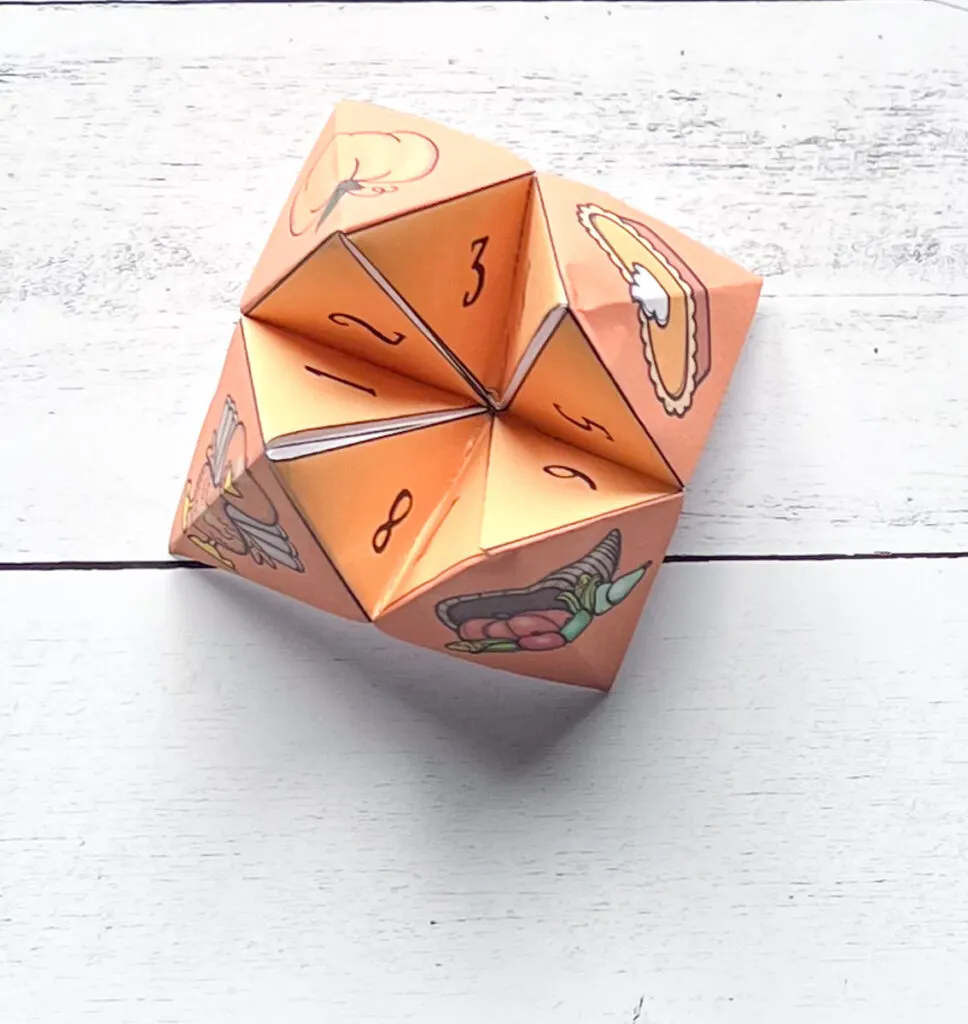 It has an assembled paper fortune teller from its outer part perfect for Thanksgiving with its orange color, placed in a black and white wood background.