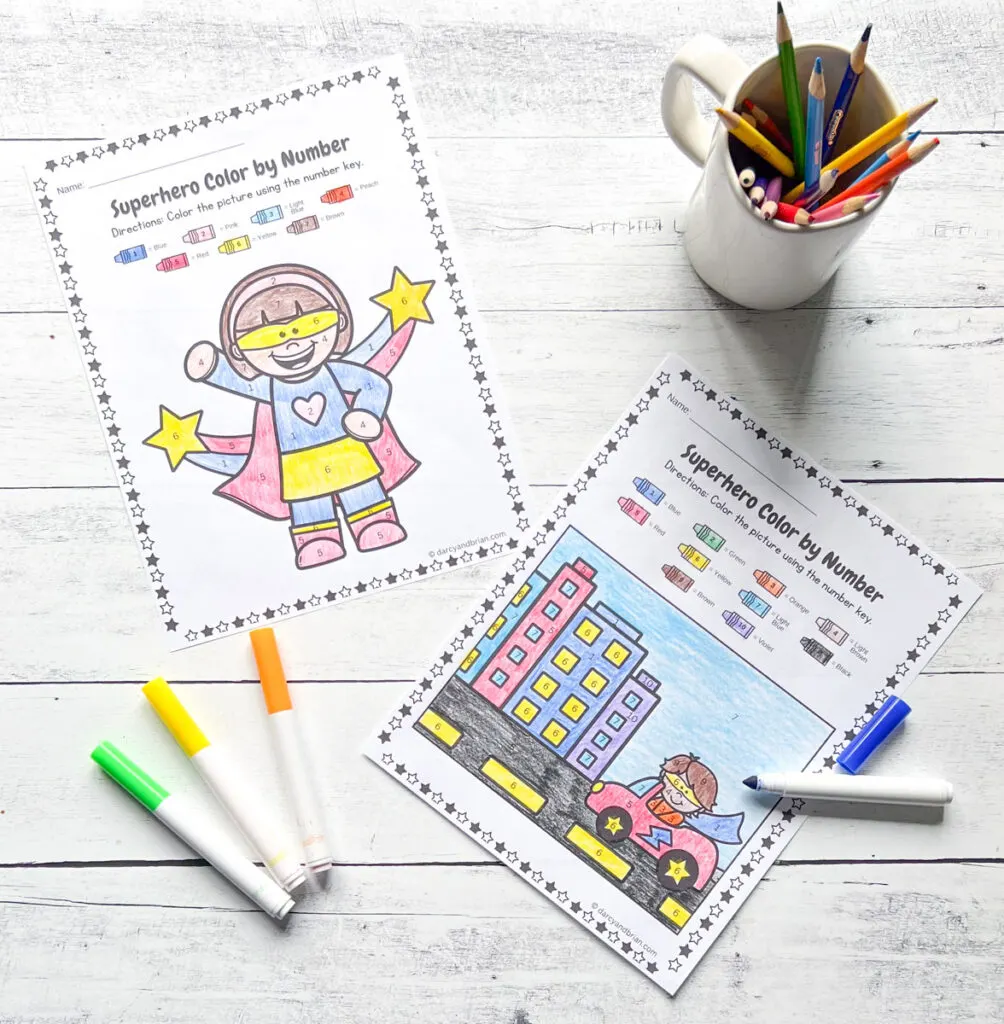 Two completed color by number worksheets with different Superhero images, there are color markers, and color pencils.