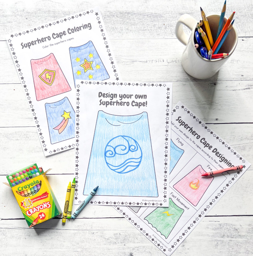 Three finished cape coloring sheets overlapping each other on a white wood table. A box of crayons is in the bottom left corner and a mug with colored pencils is in the top right.