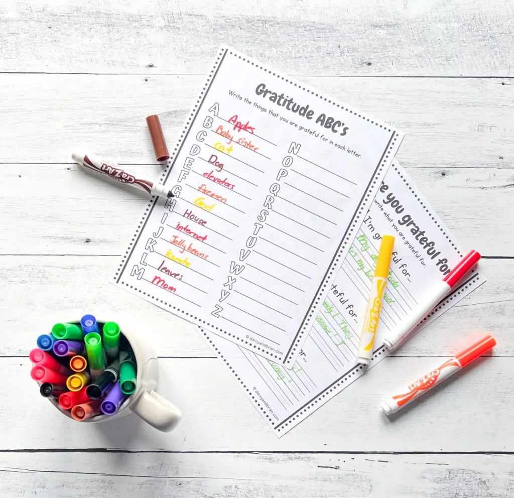 A partially filled out gratitude list for each letter of the alphabet.  A mug with markers is in the bottom left corner and markers lay next to the pages.