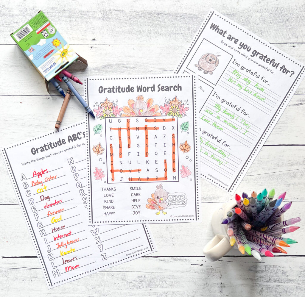 Three different gratitude activity sheets spread out on a white wood table next to crayons and markers. The top one is a word search.