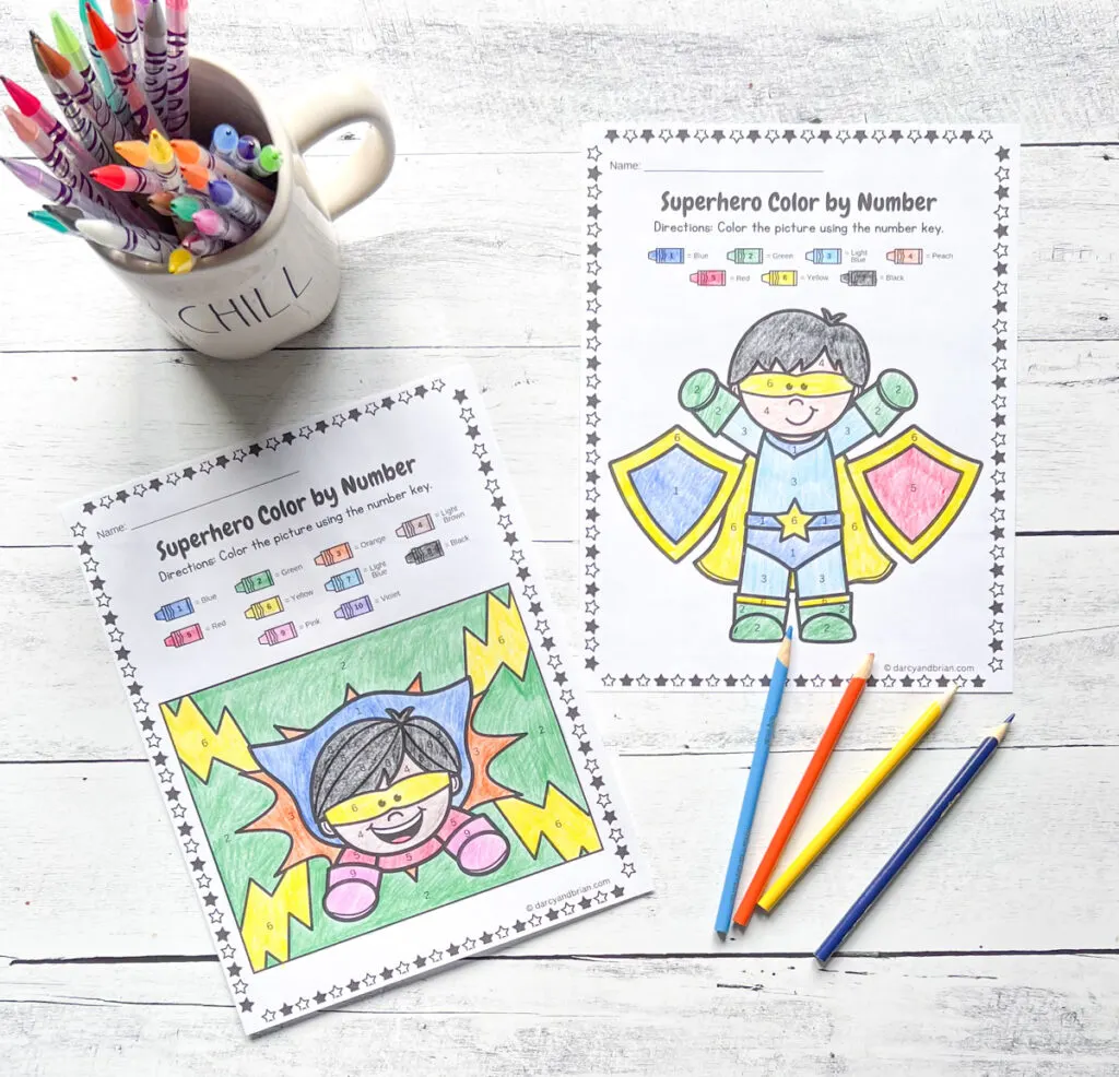 Two pages of color by number worksheets with different Superhero images, there are color pens, and color pencils, all placed in a black and white wood background.