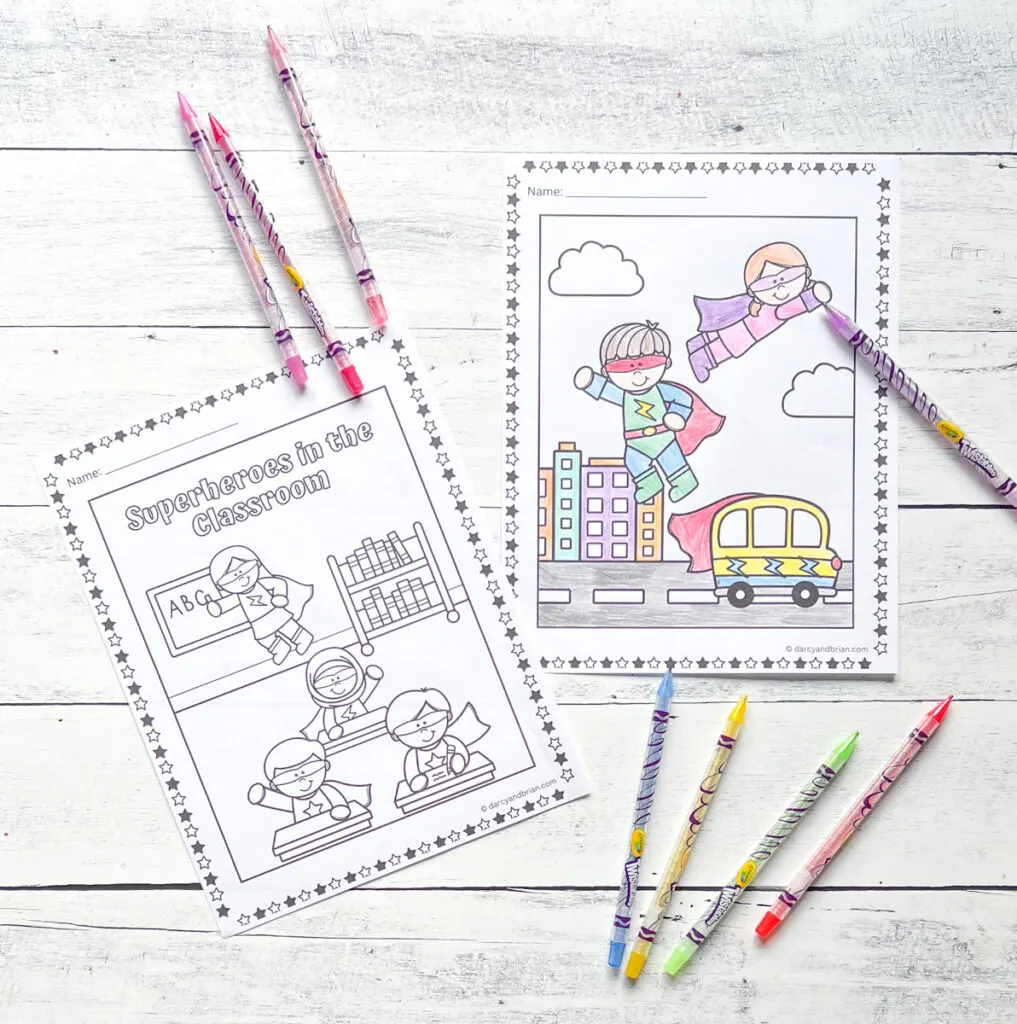 There are two pages of superhero themed coloring pages with some color pencils placed on a black and white wood background.