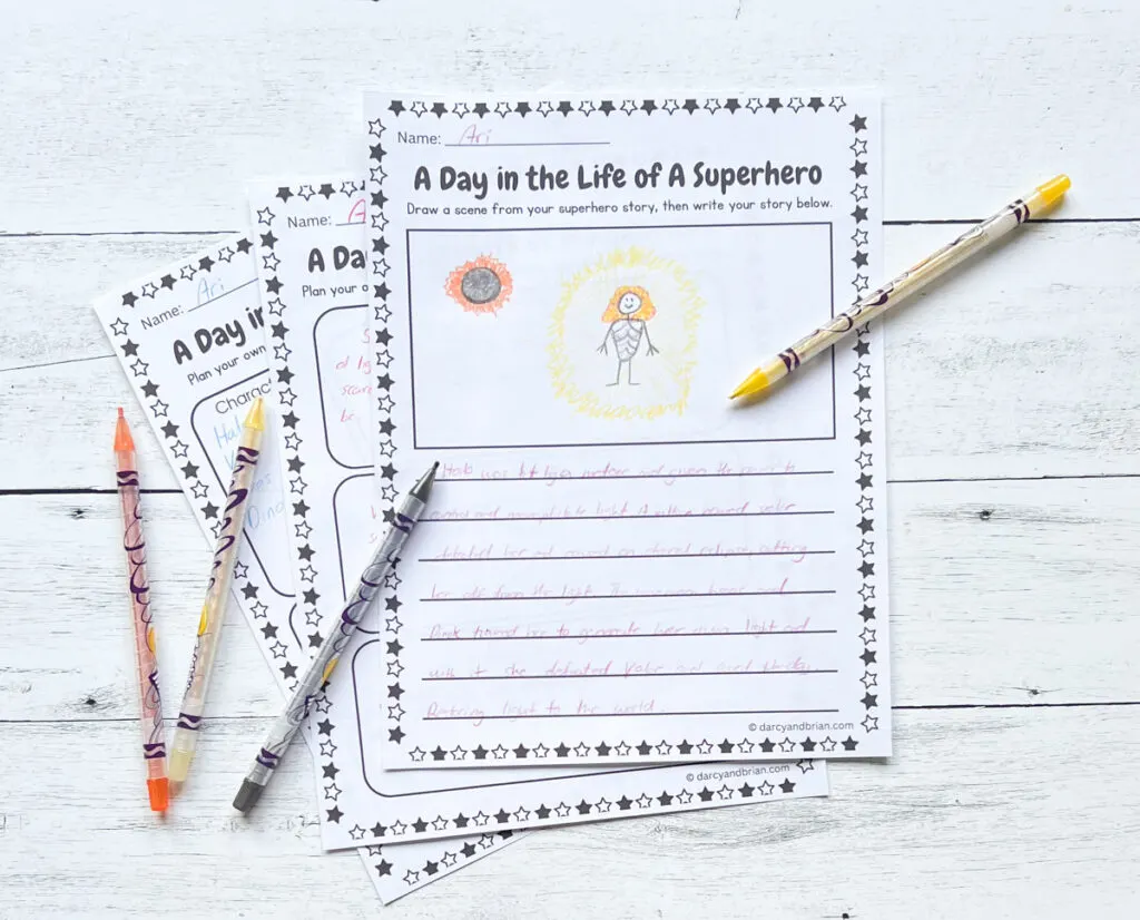 It has three pages of printables where kids can draw and write stories about spending the day as a superhero. There are some color pens placed around the papers on a black and white wood background.