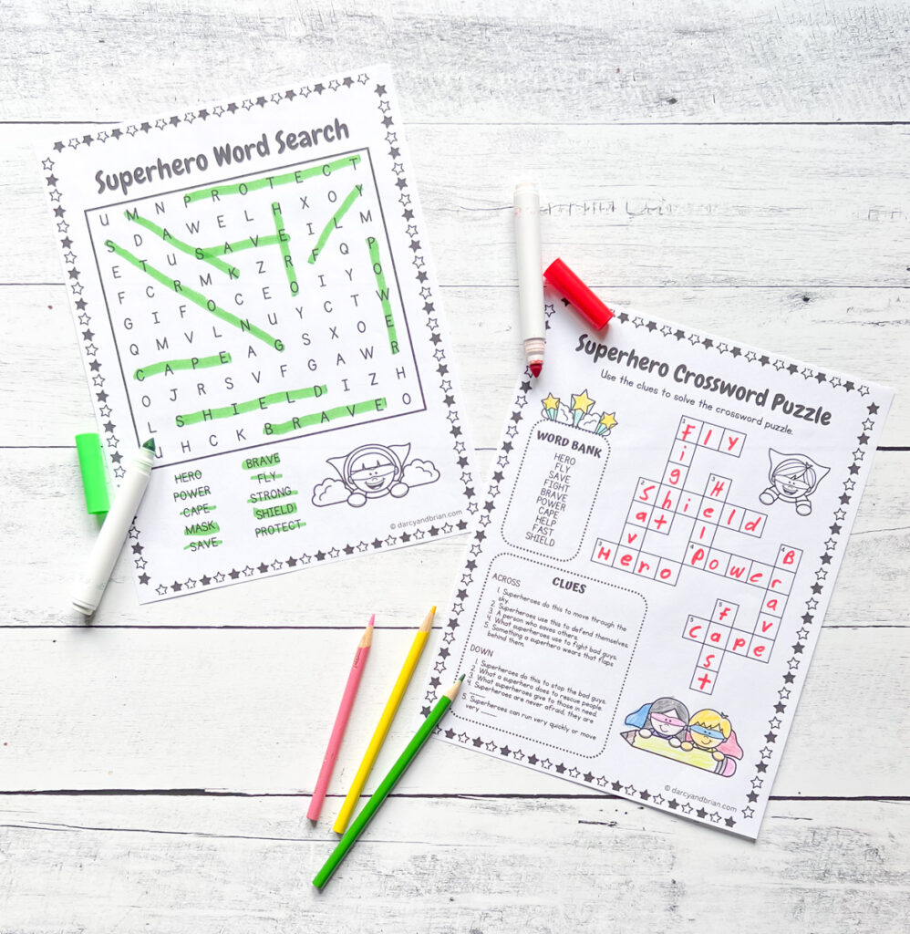 There are two pages of activities like the superhero Word Search and Crossword Puzzle with some markers and color pencils placed in a black and white wood background.