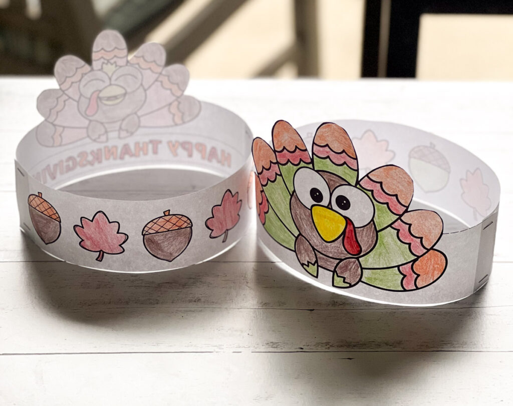 Two finished paper turkey headbands colored in are laying on a table outside. The headband has images of leaves and acorns decorating it.