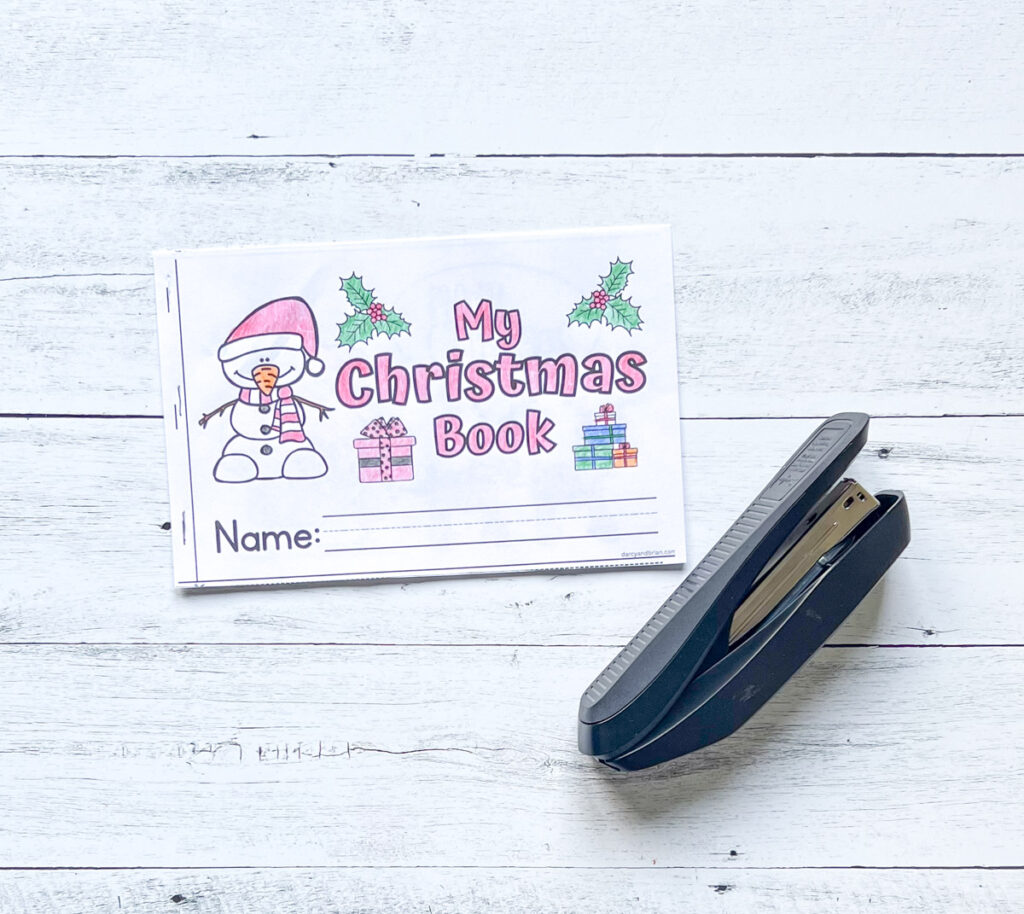 There's a cover page of the mini book Christmas-themed where kids can personalized by writing their names and design its background. There's also a stapler placed in a black and white wood background.