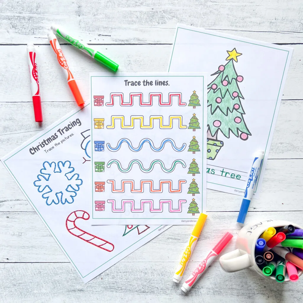 It has three pages of tracing activity with colors on the Christmas Tree, traced lines, and some Christmas decors such as snowflakes and cane, along with the markers placed in a black and white wood background.