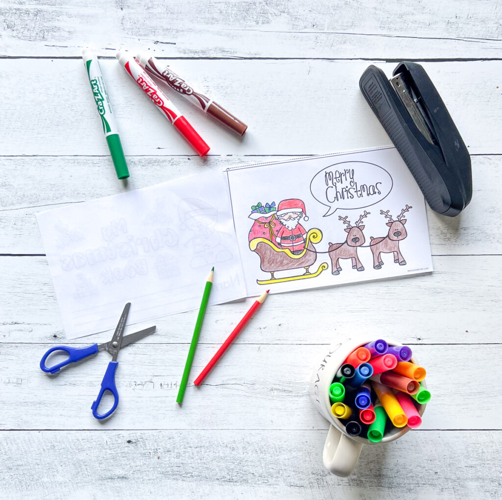 There's a cover page of mini book featuring Santa, his sleigh, and two reindeer. There are also materials such as, markers, scissors, color pencils, stapler, all placed on a black and white wood background.